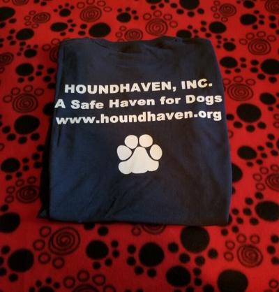 On a red background with black paw prints sits a black folded t-shirt the writing in white on the front of the t-shirt the first row says, "HOUNDHAVEN, INC. The second row says, "A Safe Have for Dogs". The third row sayd "www.houndhaven.org". The last row is a large white paw print.