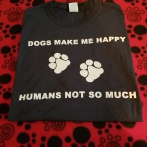 On a red background with black paw prints sits a black folded t-shirt the writing in white on the front of the t-shirt says "Dogs Make Me Happy". The next row has 2 white paw prints. The final row says "Humans Not So Much".