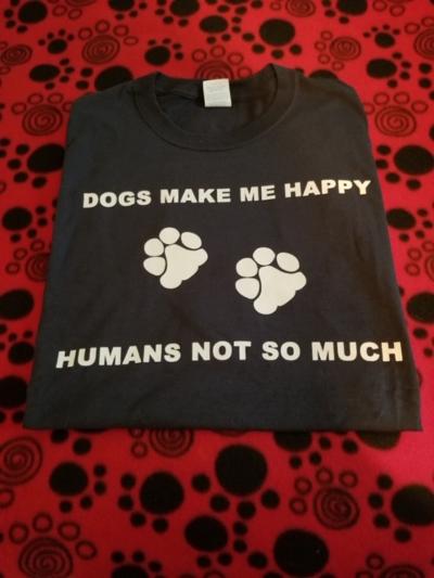 On a red background with black paw prints sits a black folded t-shirt the writing in white on the front of the t-shirt says "Dogs Make Me Happy". The next row has 2 white paw prints. The final row says "Humans Not So Much".