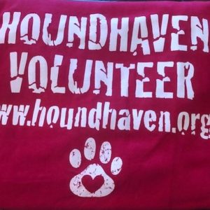 This is a picture of a red t-shirt. In white lettering on the t-shirt it reads "HOUNDHAVEN VOLUNTEER" on the first row. The next row says, "www.houndhaven.org". Below the lettering is a dog paw with a heart cut out of the pad.