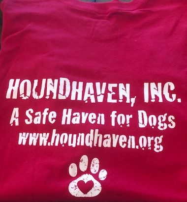 This is a picture of a red t-shirt. In white lettering on the t-shirt it reads "HOUNDHAVEN, INC" on the first row. The next row says, "A Safe Have for Dogs". The third row says, "www.houndhaven.org". Below the lettering is a dog paw with a heart cut out of the pad.